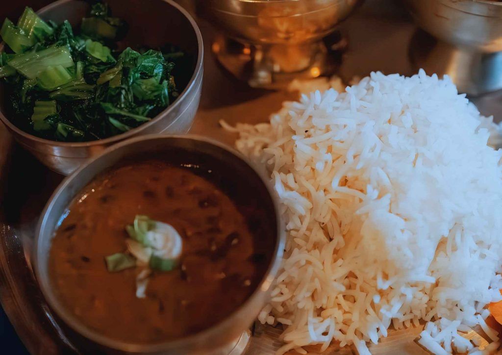 A traditional meal featuring a mound of fluffy white rice served with a bowl of lentil soup (dal) garnished with sliced green onions and a side of cooked leafy greens, arranged on a metal platter.